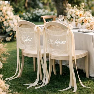 Puede incluir: Dos sillas color crema decoradas para una boda, con respaldos de encaje y largas cintas. El encaje de las sillas dice "Just Married". Las sillas est&aacute;n en un c&eacute;sped verde cerca de una mesa con arreglos florales.
