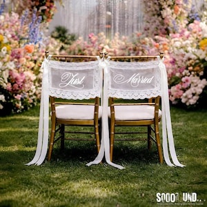 Puede incluir: Dos sillas doradas con cojines blancos decoradas para una boda. Cada silla tiene una pancarta de encaje blanco con las palabras "Just" y "Married" en escritura elegante. Largas cintas blancas cuelgan de las sillas, con un arco floral en el fondo.