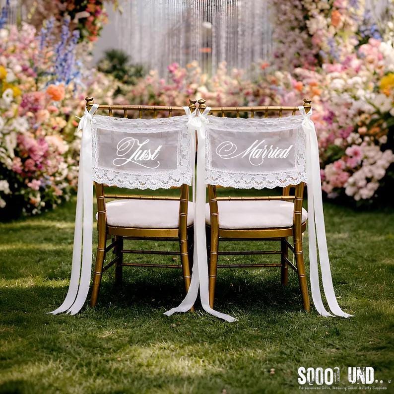 Puede incluir: Dos sillas doradas con cojines blancos decoradas para una boda. Cada silla tiene una pancarta de encaje blanco con las palabras "Just" y "Married" en escritura elegante. Largas cintas blancas cuelgan de las sillas, con un arco floral en el fondo.