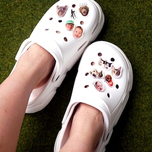 May include: White clogs adorned with decorative charms featuring images of dogs and people. The clogs are shown on a green surface. The text "SOOOO UND..." is visible in the top left corner.