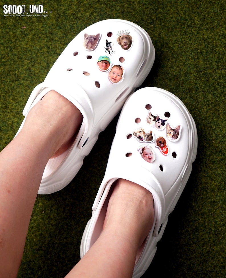 May include: White clogs adorned with decorative charms featuring images of dogs and people. The clogs are shown on a green surface. The text "SOOOO UND..." is visible in the top left corner.