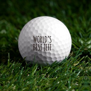 May include: A white golf ball with the text "WORLD'S BEST JEFF" printed on it. The golf ball is sitting on green artificial grass.