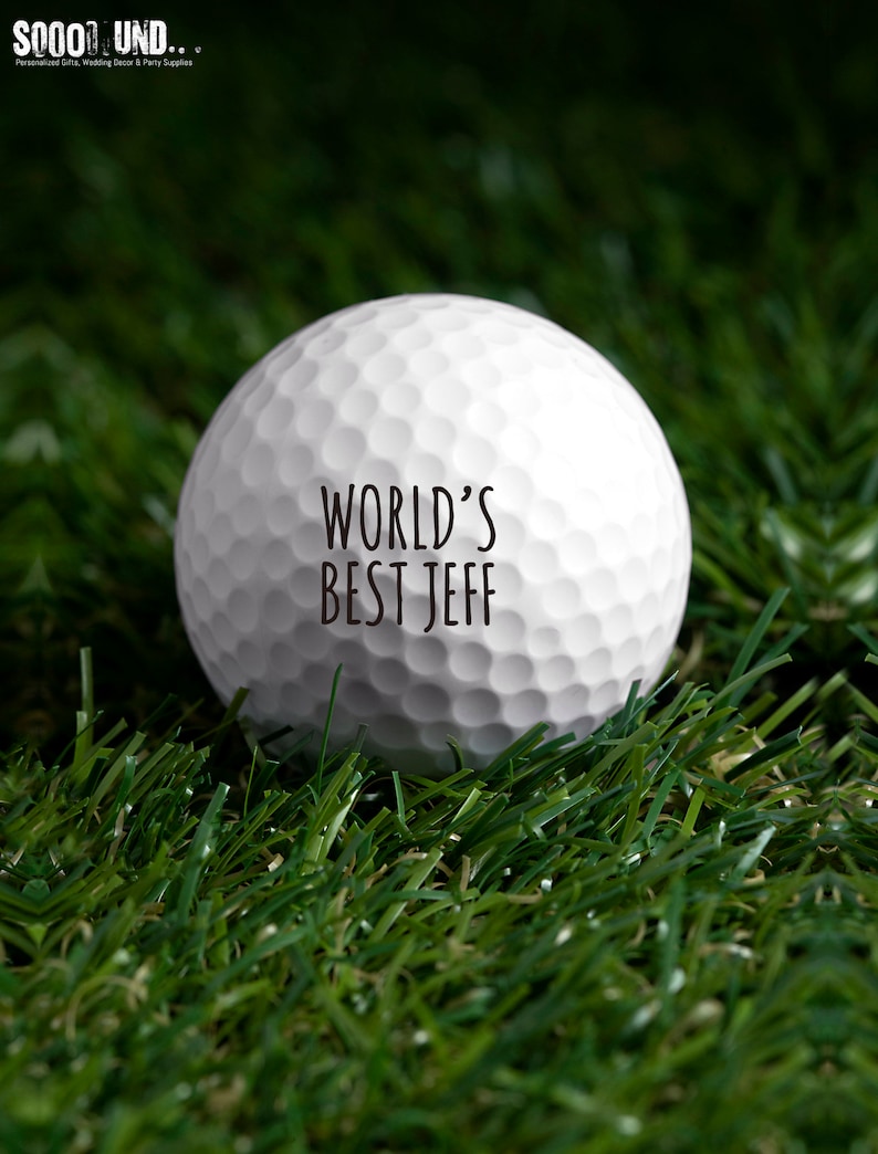 May include: A white golf ball with the text "WORLD'S BEST JEFF" printed on it. The golf ball is sitting on green artificial grass.
