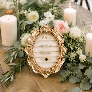 May include: An oval-shaped gold ring display box with an ornate, vintage-style frame. The interior features a cream-coloured velvet insert with three horizontal slots holding three rings. The letters B and L with a heart are printed at the top.