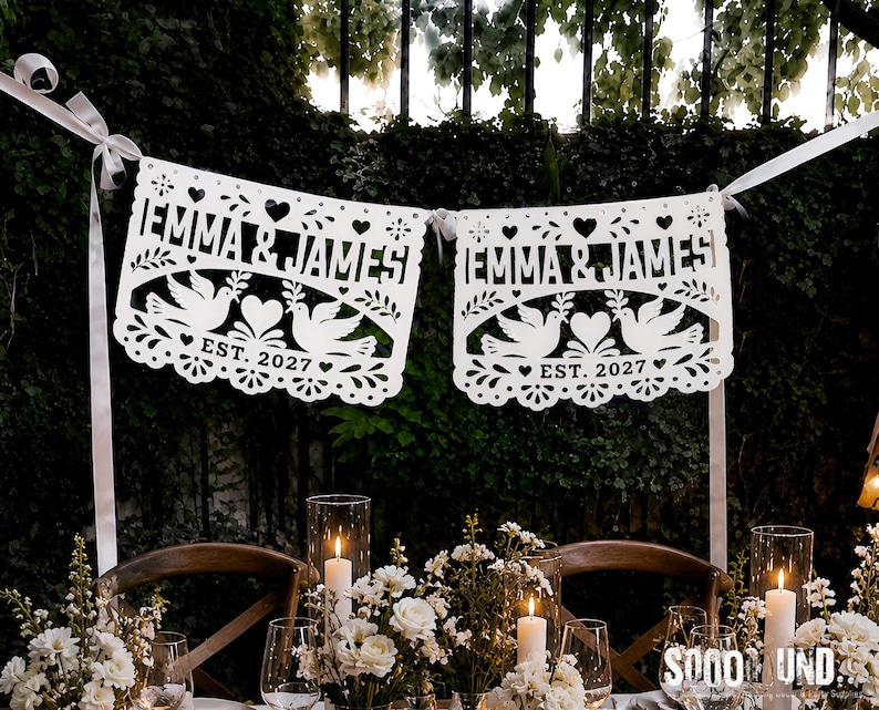 May include: A white paper banner featuring two rectangular panels with intricate cut-out designs. Each panel displays the text EMMA & JAMES, two doves holding olive branches, a central heart, and the text EST. 2027, all suspended by a white ribbon.