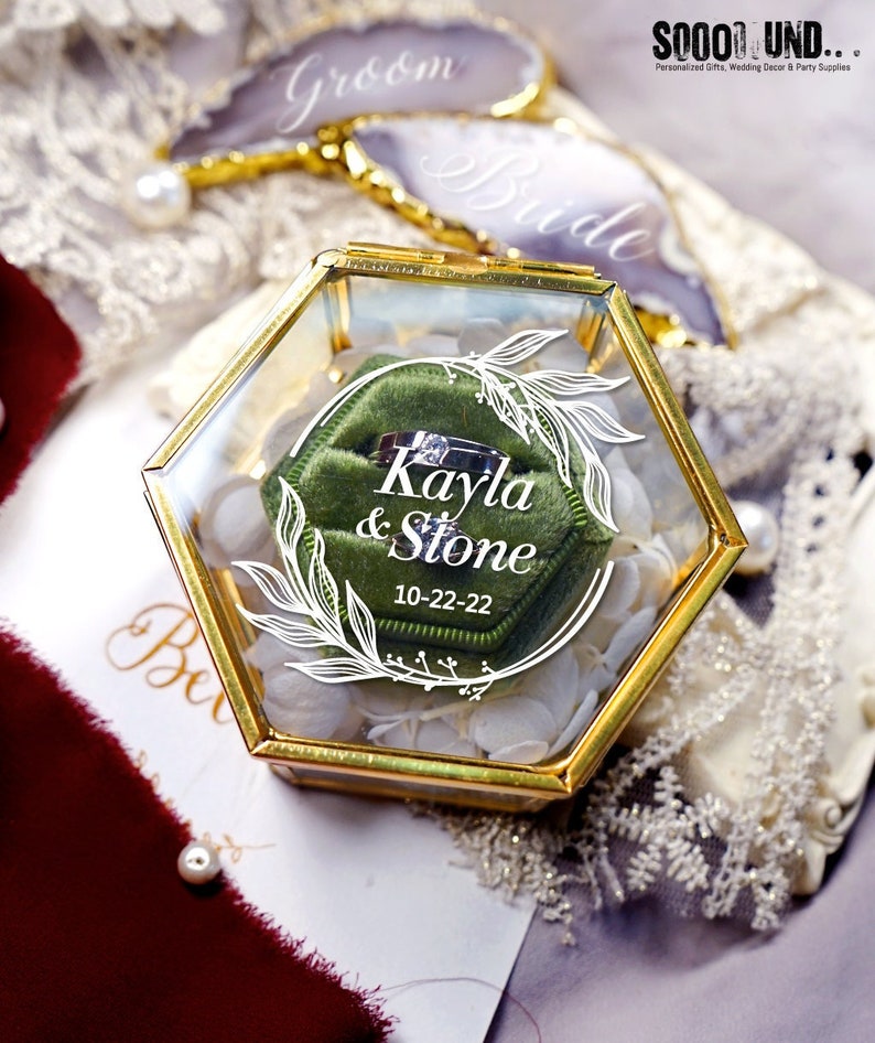 May include: A hexagonal glass ring box with a gold trim. The box has a green velvet cushion inside with a ring on it. The box is decorated with a white floral design and the text "Kayla & Stone 10-22-22".