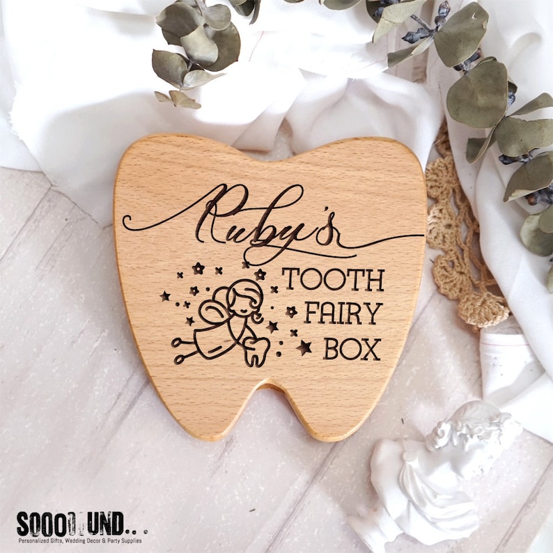 Puede incluir: Una caja de madera con forma de diente con la inscripci&oacute;n "Ruby's Tooth Fairy Box" y una peque&ntilde;a ilustraci&oacute;n de un hada sosteniendo un diente.