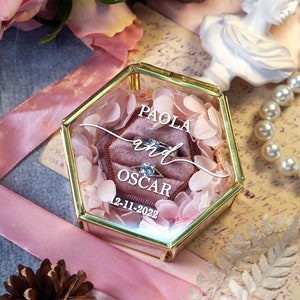 May include: A hexagonal glass ring box with a gold trim and a pink velvet interior. The box is filled with pink flower petals and contains two rings. The box is personalized with the names "PAOLA and OSCAR" and the date "12-11-2022".