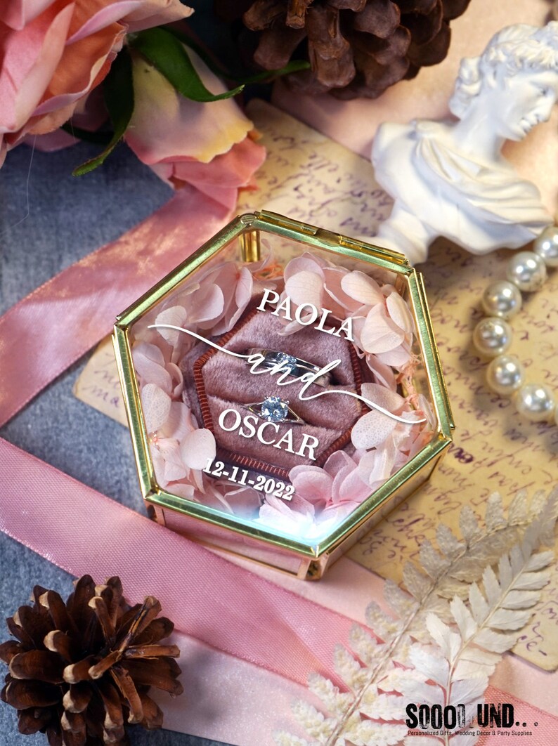 May include: A hexagonal glass ring box with a gold trim and a pink velvet interior. The box is filled with pink flower petals and contains two rings. The box is personalized with the names "PAOLA and OSCAR" and the date "12-11-2022".