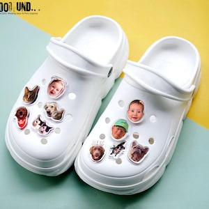 May include: White clogs decorated with various photo charms of pets and people. The charms are clear and feature images of dogs, cats, and a baby. The clogs are on a yellow and blue background. The text "SOOOOL UND..." is visible in the top left corner.