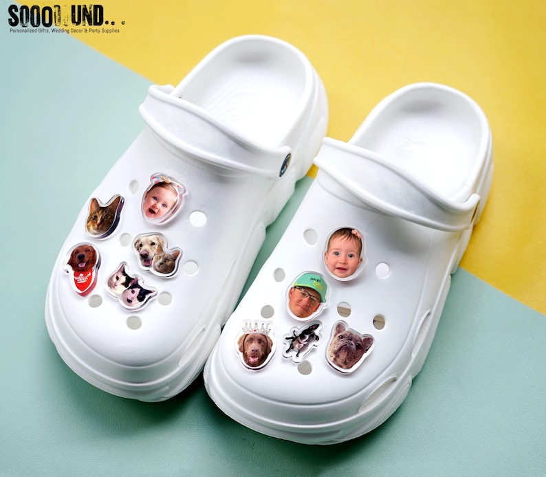 May include: White clogs decorated with various photo charms of pets and people. The charms are clear and feature images of dogs, cats, and a baby. The clogs are on a yellow and blue background. The text "SOOOOL UND..." is visible in the top left corner.