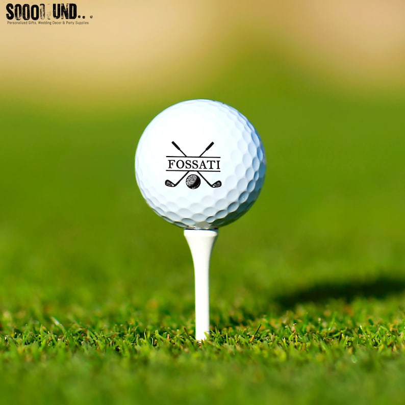 May include: A white golf ball with a black design featuring crossed golf clubs and the name "FOSSATI" in a banner.