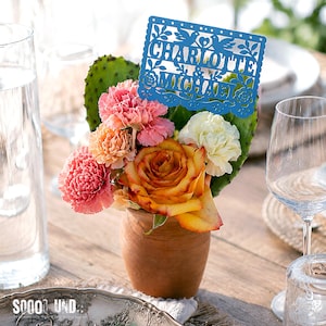 May include: A terracotta pot containing a floral arrangement of pink and orange carnations, a yellow and orange rose, and a white carnation, accented by a green prickly pear cactus pad and a blue laser-cut sign reading CHARLOTTE & MICHAEL.