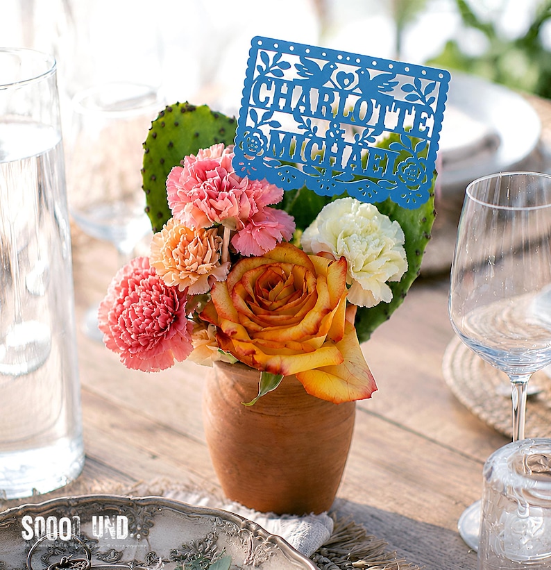 May include: A terracotta pot containing a floral arrangement of pink and orange carnations, a yellow and orange rose, and a white carnation, accented by a green prickly pear cactus pad and a blue laser-cut sign reading CHARLOTTE & MICHAEL.