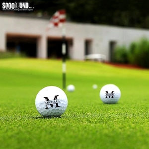 May include: Two white golf balls with black lettering. The text on the balls reads "M" and "MICHEAL". The balls are on a green golf course.