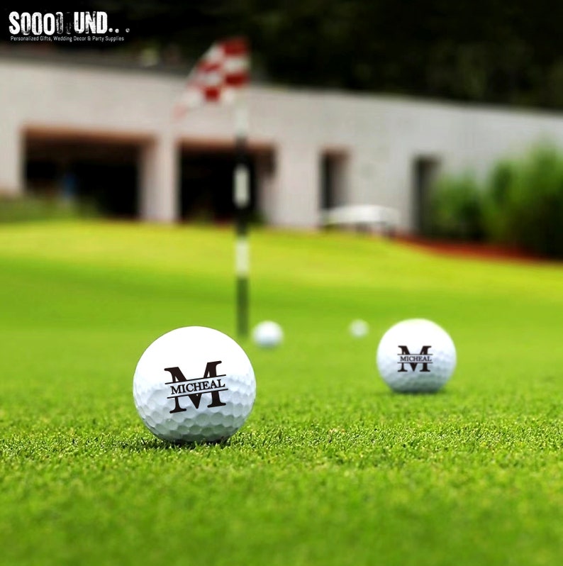 May include: Two white golf balls with black lettering. The text on the balls reads "M" and "MICHEAL". The balls are on a green golf course.