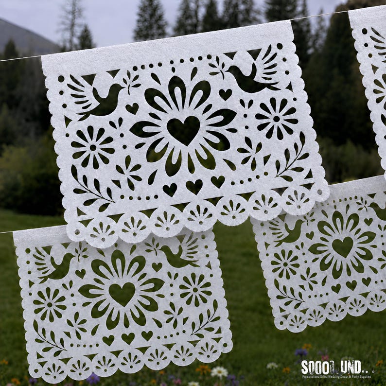 May include: A white paper banner featuring intricate cut-out designs of central flowers, hearts, and birds holding olive branches. The rectangular panels have scalloped edges and are strung together, displaying a repeating pattern of floral motifs.
