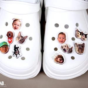 May include: Two white clogs decorated with various photo charms. One clog features a baby's face, a dog, a cat, and a person wearing a hat. The other clog has a baby's face, dogs, and a bulldog.