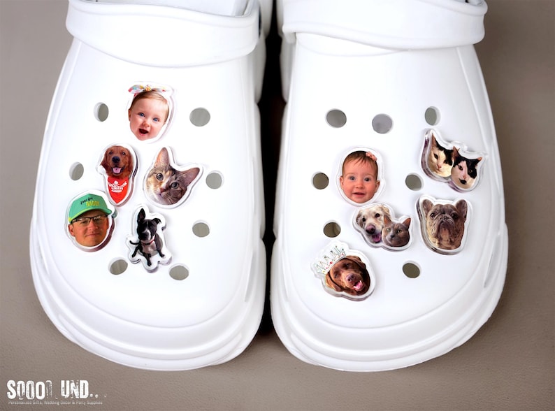 May include: Two white clogs decorated with various photo charms. One clog features a baby's face, a dog, a cat, and a person wearing a hat. The other clog has a baby's face, dogs, and a bulldog.