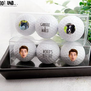 May include: Four white golf balls with personalized images and text. The golf balls are in a clear plastic box. The text on the golf balls reads "Gabriele & Marco" and "World's Best Jeff".