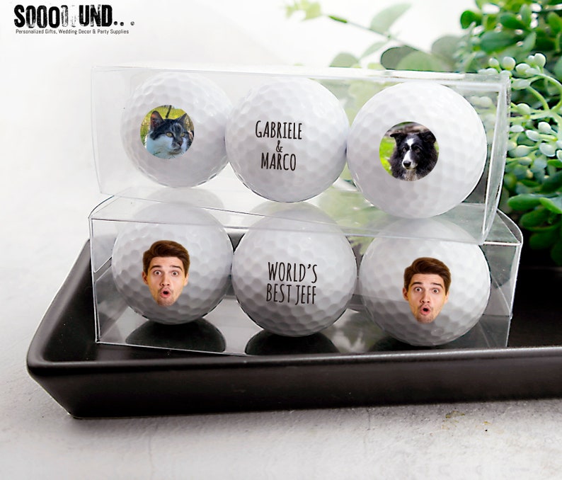 May include: Four white golf balls with personalized images and text. The golf balls are in a clear plastic box. The text on the golf balls reads "Gabriele & Marco" and "World's Best Jeff".