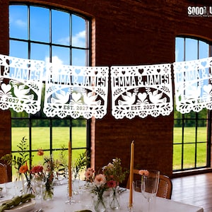 May include: A white paper banner featuring intricate cut-out designs of two doves facing each other with a heart in the center. The text "EMMA & JAMES" and "EST. 2027" is clearly visible on each panel, surrounded by decorative floral and heart patterns.