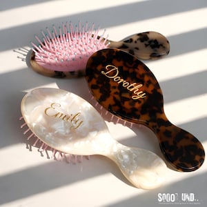 May include: Three personalized hairbrushes with different patterns and colors. One is pink with blue bristles, another is tortoiseshell with the name "Dorothy" in gold, and the third is pearlescent with "Emily" in gold. The brushes are oval-shaped with a handle.