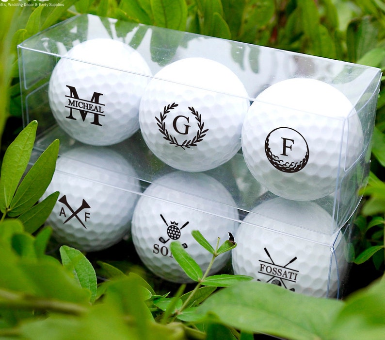 May include: Six white golf balls in a clear plastic box. Each ball has a different personalized design, including initials, names, and golf-themed graphics.