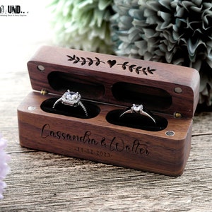 K&ouml;nnte beinhalten: Eine Holzkiste f&uuml;r Ringe mit zwei F&auml;chern, graviert mit den Namen "Cassandra & Walter" und dem Datum "11-12-2023". Die Box hat ein Herz- und Bl&auml;tterdesign auf der Oberseite.