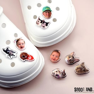 May include: White clogs adorned with various photo charms, including faces of a baby, a man wearing a green hat, and several cats and dogs. The charms are clear and have a glossy finish. The clogs are on a light pink surface.
