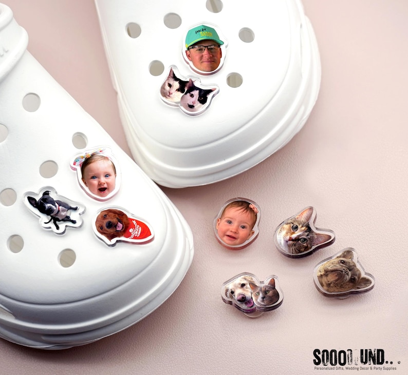 May include: White clogs adorned with various photo charms, including faces of a baby, a man wearing a green hat, and several cats and dogs. The charms are clear and have a glossy finish. The clogs are on a light pink surface.