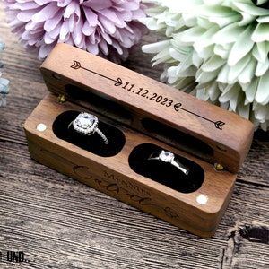 K&ouml;nnte beinhalten: Eine Holzkiste f&uuml;r Ringe mit einem Scharnierdeckel. Die Kiste ist mit dem Text "Mr & Mrs" und "11.12.2023" graviert. Die Kiste hat zwei F&auml;cher, jedes mit einem Samtbezug, und ist f&uuml;r zwei Ringe ausgelegt.