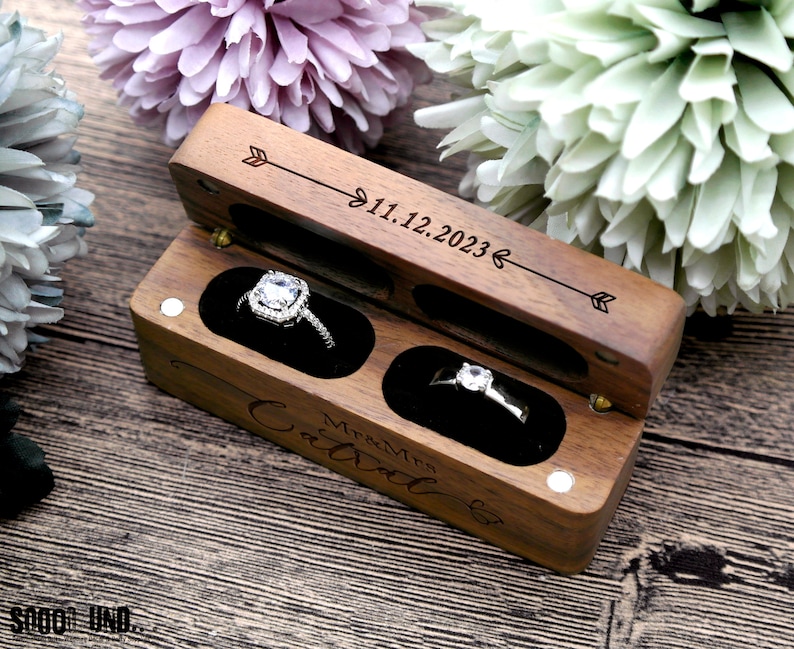 K&ouml;nnte beinhalten: Eine Holzkiste f&uuml;r Ringe mit einem Scharnierdeckel. Die Kiste ist mit dem Text "Mr & Mrs" und "11.12.2023" graviert. Die Kiste hat zwei F&auml;cher, jedes mit einem Samtbezug, und ist f&uuml;r zwei Ringe ausgelegt.