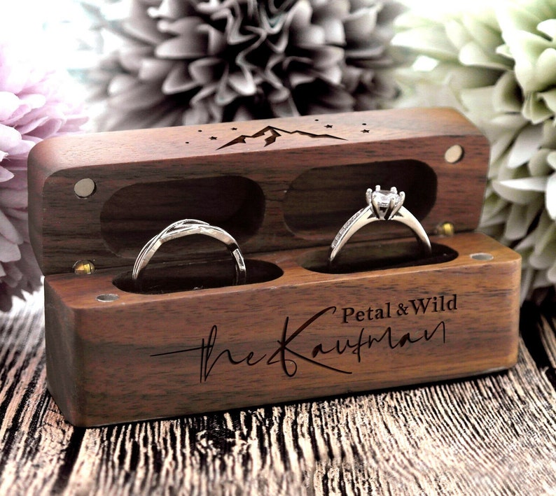 K&ouml;nnte beinhalten: Eine Holzkiste f&uuml;r Ringe mit einem Berg- und Sternenmotiv. Die Box hat zwei F&auml;cher f&uuml;r Ringe und ist mit "Petal & Wild the Kaufman" graviert.