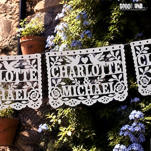 May include: A white paper banner featuring intricate laser-cut designs of two birds, a heart, and floral patterns. The text "CHARLOTTE & MICHAEL" is prominently displayed in the center. The banner is suspended, showcasing delicate cutouts.