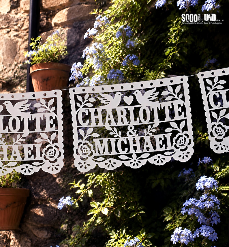 May include: A white paper banner featuring intricate laser-cut designs of two birds, a heart, and floral patterns. The text "CHARLOTTE & MICHAEL" is prominently displayed in the center. The banner is suspended, showcasing delicate cutouts.