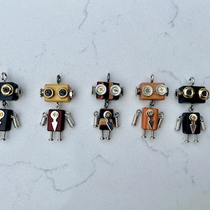 May include: Five wooden robot charms with different wood grain patterns. Each charm has a black, gold, and silver metal accents. The charms are hanging from a white surface.