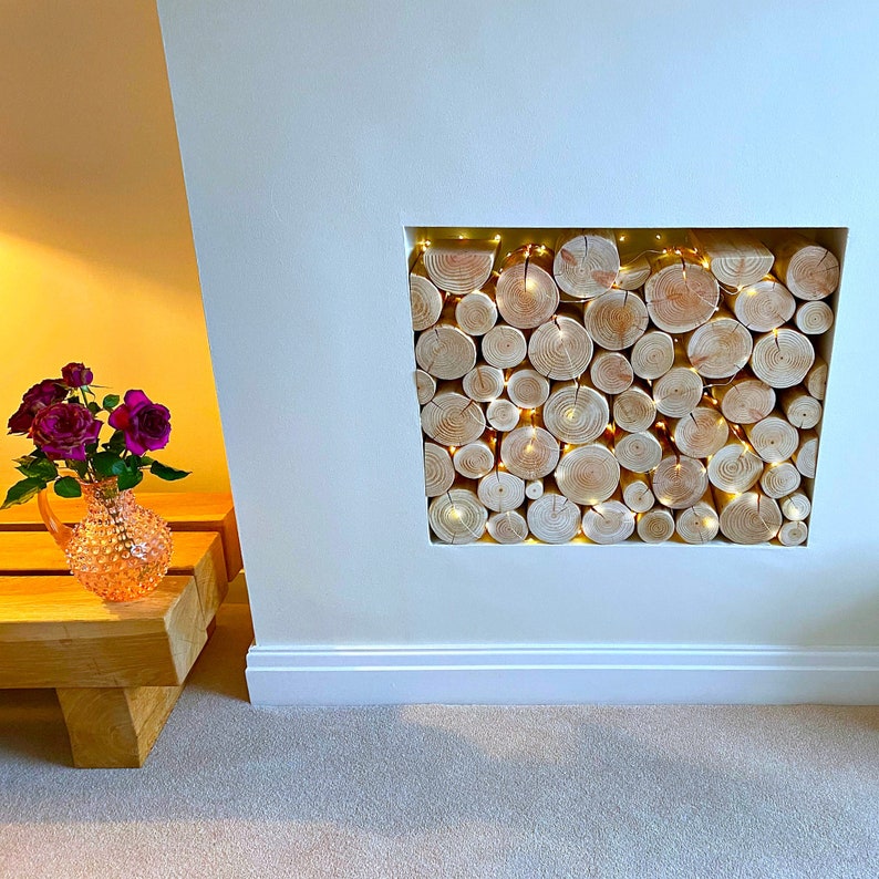 Neat round clean real wood decorative logs from The Log Basket for interior display fill a fireplace alcove stacked to show the textural growth rings or end grains with added warmth and sparkle of tiny LED fairy lights wound through the logs.