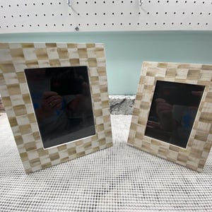May include: Two square picture frames featuring a checkered pattern in alternating shades of light beige and white. Each frame has a wide border with a smooth finish and a central rectangular opening for displaying photographs or artwork.