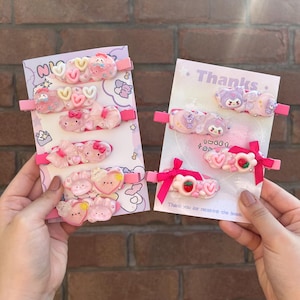 Peut inclure: Deux ensembles de pinces à cheveux roses et blanches avec des personnages de dessins animés et des cœurs. Les pinces sont sur un fond rose et blanc avec le texte "Merci" et "Douces Rêves".