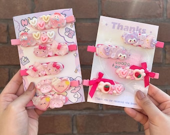 Può includere: Due set di fermagli per capelli rosa e bianchi con personaggi dei cartoni animati e cuori. I fermagli sono su uno sfondo rosa e bianco con il testo "Grazie" e "Dolci Sogni".