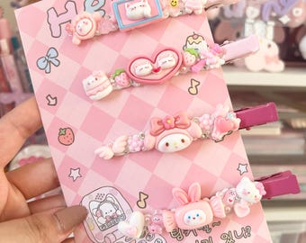 Può includere: Set di cinque mollette per capelli rosa e bianche con vari personaggi dei cartoni animati e disegni. Le mollette presentano un personaggio di Hello Kitty, un cuore con due gatti, un telefono con un gatto, un cupcake con un gatto e una fragola con un gatto. Le mollette sono su uno sfondo rosa e bianco con il testo "Hello" e "강아지 있니?" che si traduce in "Hai un cane?"