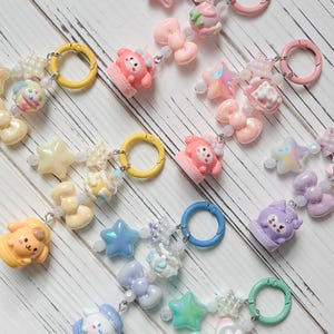 Peut inclure: Plusieurs bracelets à breloques colorés avec divers éléments décoratifs. Les breloques comprennent des étoiles, des nœuds, des ours en peluche et d'autres motifs mignons. Les bracelets comportent des fermoirs circulaires jaunes, roses, bleus et verts, et sont enfilés avec de petites perles.
