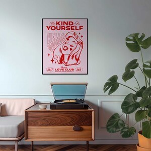 May include: A pink and red poster with the text "Be Kind to Yourself" and "Hello Gorgeous" in a retro font. The poster also features a cartoon woman with a heart-shaped beauty mark and the text "The Self Love Club 24/7 365 Because You Are Worthy".
