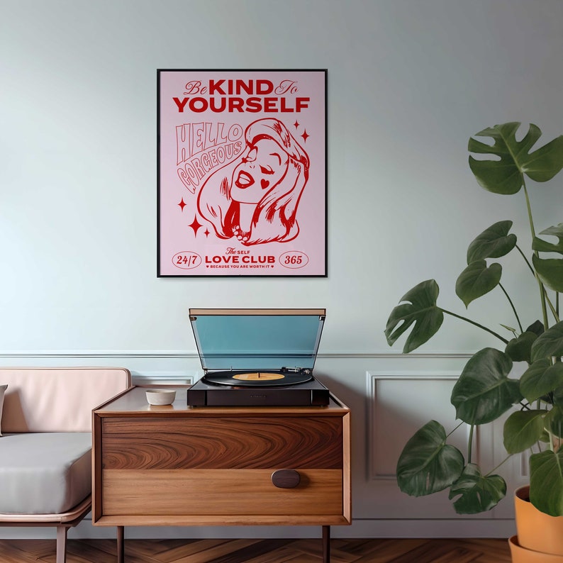May include: A pink and red poster with the text "Be Kind to Yourself" and "Hello Gorgeous" in a retro font. The poster also features a cartoon woman with a heart-shaped beauty mark and the text "The Self Love Club 24/7 365 Because You Are Worthy".