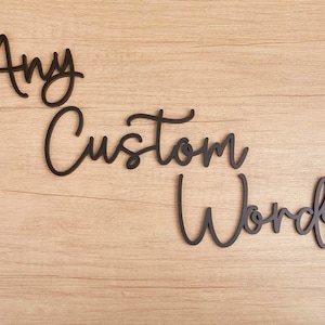 Può includere: Scritte ritagliate in nero che compongono le parole Any Custom Words in un carattere corsivo fluido. Le lettere sono disposte su tre righe separate, caratterizzate da bordi lisci e una finitura opaca scura che risalta sul legno chiaro.