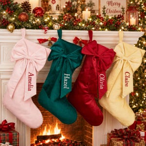 Könnte beinhalten: Vier Weihnachtssocken aus Samt hängen in einer Reihe, jede mit einer großen dekorativen Schleife an der Oberseite. Die Socken sind hellrosa, dunkelgrün, tiefrot und blassgelb und mit den Namen Amelia, Hazel, Olivia und Claire bestickt.