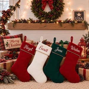 May include: Four knitted Christmas stockings displayed in a row, consisting of two red, one white, and one dark green stocking. Each stocking features a folded cuff with gold-coloured trim and a name embroidered in cursive script: Hazel, Everett, June, and Weston.