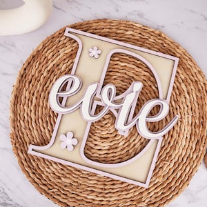 Könnte beinhalten: Ein Holzschild mit dem Namen "Evie", der über einem großen Buchstaben "E" liegt. Der Buchstabe und der Name sind in Weiß und Hellrosa gehalten, mit kleinen Blumenakzenten. Das Schild liegt auf einer gewebten, runden Matte.