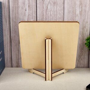 Puede incluir: Un marco de fotos de madera con un acabado natural, sostenido por un caballete de madera. El marco es cuadrado con esquinas redondeadas. El soporte tiene una base triangular y un soporte vertical. Un libro azul y una peque&ntilde;a planta en maceta est&aacute;n en el fondo.